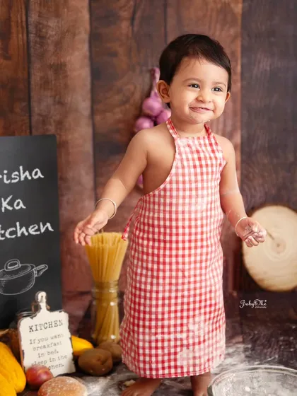 A proud little chef standing tall in his kitchen. This shot shows off the adorable checkered apron and the whole setup, from the pasta jars to the 'Prisha Ka Kitchen' sign.