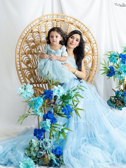 A mother and daughter looking like royalty on a beautiful peacock chair. The flowing blue gowns and floral decorations create a magical, ethereal scene.