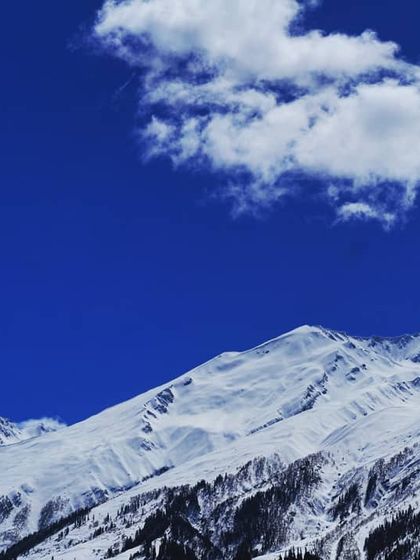 A vast expanse of snow covered mountains under a brilliant, deep blue sky. The stark contrast and the single wisp of a cloud highlight the sheer scale and purity of the high Himalayas.