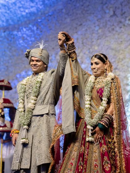 A triumphant moment for the newlyweds. The way their outfits work together on stage shows the power of complementary styling. His subtle green and her rich reds and pinks create a harmonious visual.
