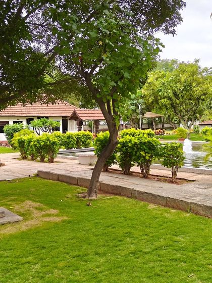 A view across the lawn towards the pond and a heritage building, with a traditional stone lamp (Deepastambham) in the foreground.