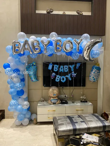 A clear view of the 'Baby Boy' balloon arch over a TV, featuring silver foil letters, a moon-shaped foil balloon, and other themed elements.