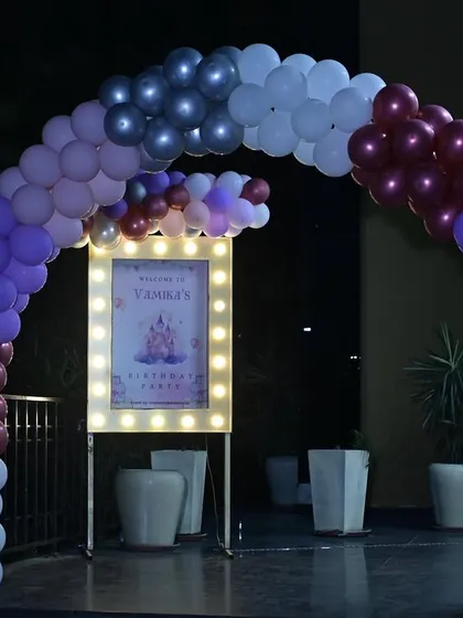 The entrance to Vamika's party, with a balloon arch in shades of purple, pink, and silver, and a lit-up welcome sign.