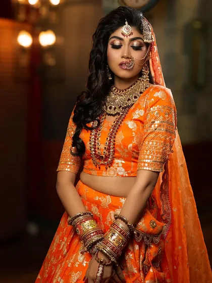 A full-length portrait showcasing a vibrant orange Banarasi lehenga. The makeup features a dramatic smokey eye and bold jewelry, creating a stunning look for a Sangeet or wedding ceremony.