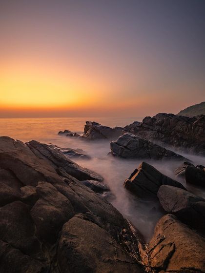A long exposure shot taken during the golden hour, where the receding tide leaves a soft, misty effect over the rocks. The warm light creates a peaceful and serene mood.