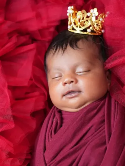 A close-up of the crowned newborn, focusing on their peaceful, sleeping face. The soft lighting highlights their delicate features against the vibrant red backdrop.