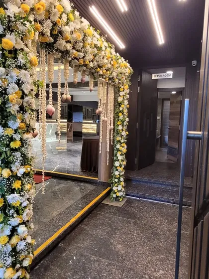 An elegant floral archway at the entrance of a banquet hall. The mix of white and yellow flowers with hanging pearl strings creates a luxurious and welcoming passage for guests.