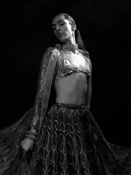 A dramatic black and white image focusing on the flow and pattern of a traditional-style garment. The model's pose and the high-contrast lighting create a powerful, artistic statement.