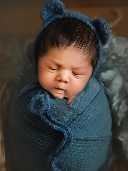 A sleeping 10-day-old newborn is photographed wearing a cute blue bonnet with bear ears. The focus on the baby's peaceful face and the soft texture of the wrap creates a gentle and heartwarming portrait.