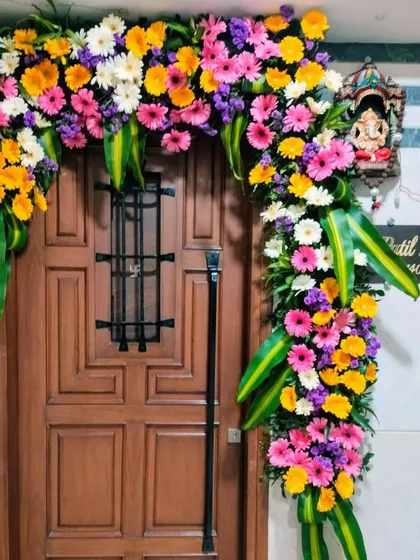 A vibrant and joyful door decoration for a wedding home. The arch is filled with a colorful assortment of gerbera daisies in pink, yellow, and orange, accented with large green leaves.