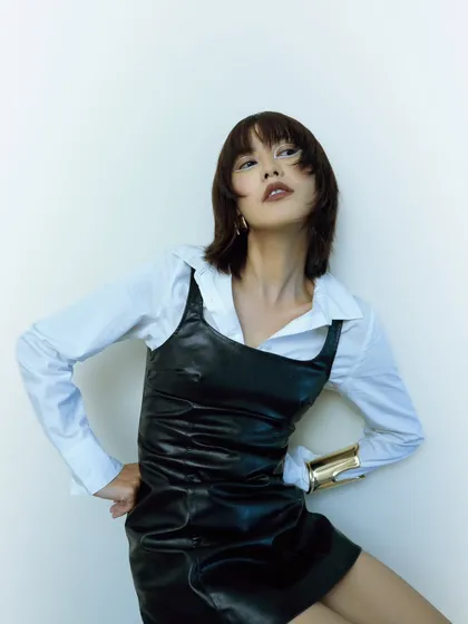 A high-energy, dynamic pose from a test shoot. The layered styling of a leather dress over a white shirt is showcased with a modern, edgy attitude.
