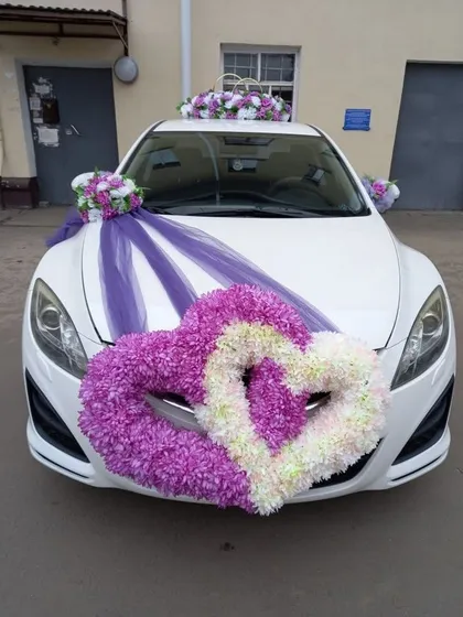 A playful and romantic car decoration featuring two interlocking hearts made of purple and white flowers. Purple ribbons and smaller bouquets on the roof and mirrors complete this lovely design.