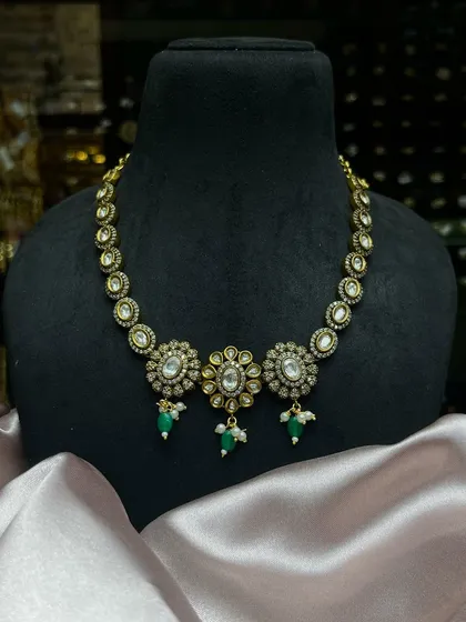 This beautiful Kundan choker features three floral motifs with green drops and pearls. It is a classic design that adds understated sparkle and sophistication to any ethnic outfit.
