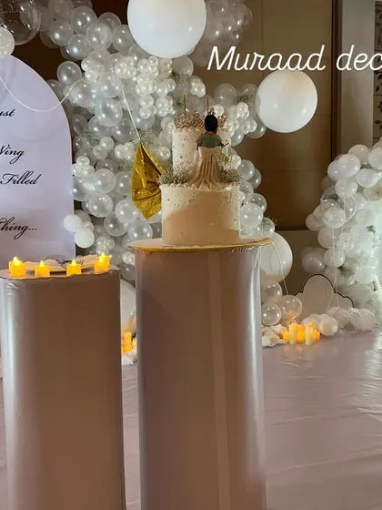 The cake-cutting area for the royal-themed party. The backdrop has a lovely quote, and the cake stands on a pedestal surrounded by white balloons that look like clouds and soft, warm lights.