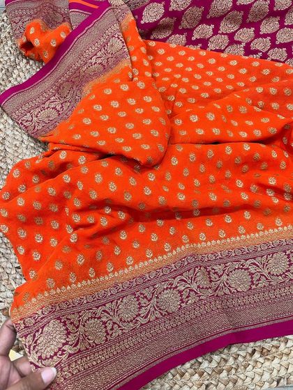 A striking orange and pink pure Banarasi Kadi georgette saree. The body is covered in delicate butis, complemented by a richly woven floral border.