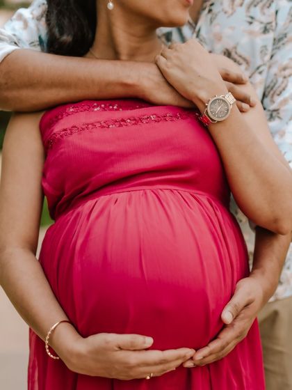 A vibrant close-up of a dad's hands embracing his partner's baby bump. The bright red dress and the detail of his watch create a modern and loving maternity image.
