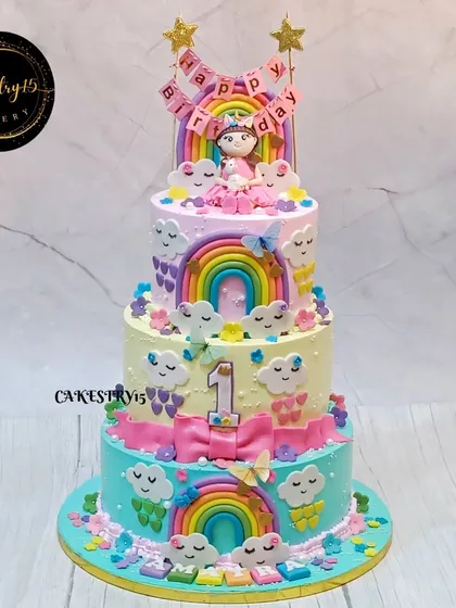 A towering four-tier rainbow-themed cake created for a truly spectacular first birthday. Each tier is a burst of color, decorated with fondant rainbows, happy clouds, and a sweet baby girl figurine on top.