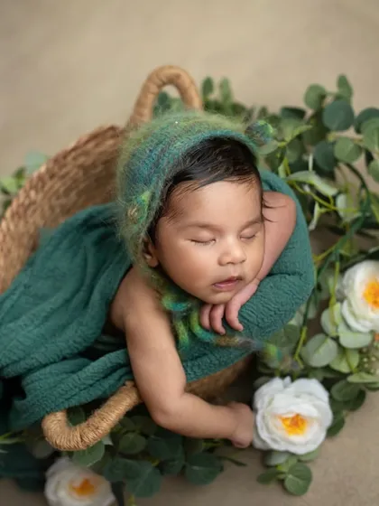 A newborn rests peacefully in a basket, wearing a fuzzy green bonnet. The textures of the bonnet and the surrounding greenery add a touch of nature to this sweet portrait.