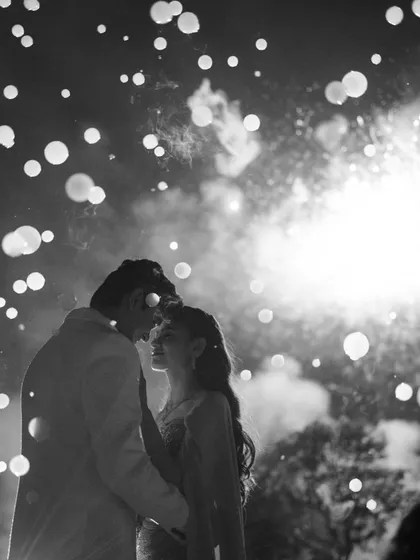 A magical sangeet moment captured in timeless black and white. The silhouette against the fireworks creates a dreamy, star-struck atmosphere, freezing a fleeting second of pure celebration and romance into an unforgettable memory.