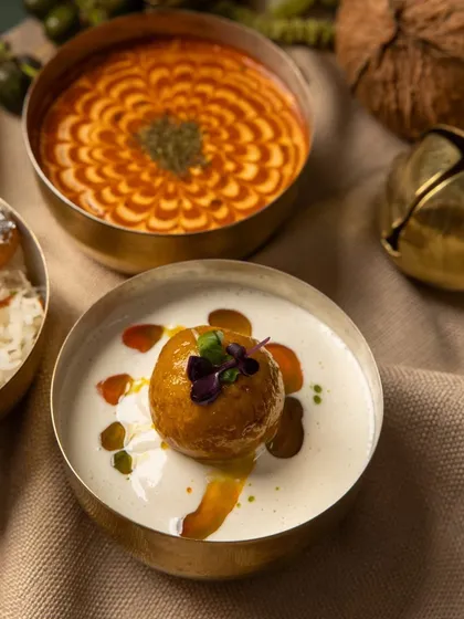 A modern Indian dessert or starter featuring a fried dumpling in a creamy white sauce, artistically garnished with oils and an edible flower.