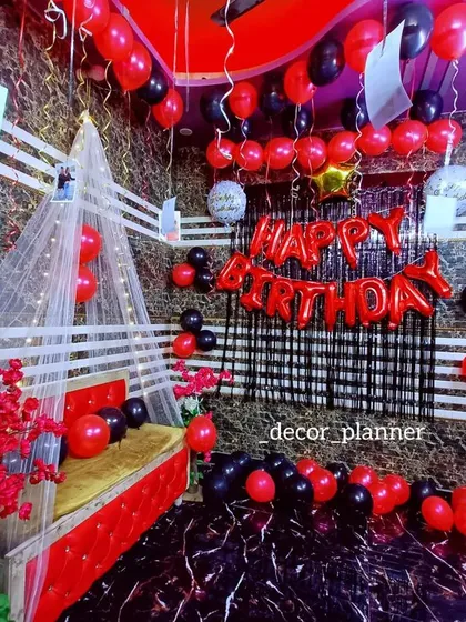 This romantic room setup features a canopy bed draped with netting, red and black balloons, and a floor scattered with heart-shaped petals. It's a classic and passionate way to say 'I love you' on a special day.