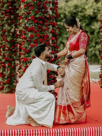 A tender and symbolic moment during a South Indian wedding, where the groom puts the toe ring on the bride, set against a stunning backdrop of red roses.