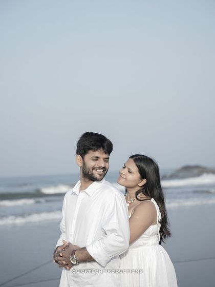 An intimate and happy portrait of a couple on a beach in Goa, with the sea and rocks in the background.