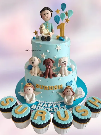 A heavenly-themed first birthday cake surrounded by matching cupcakes. The design includes the birthday boy's favorite dogs and little angel figures.