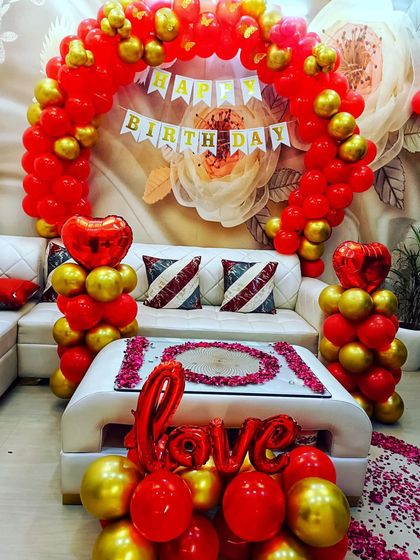 A vibrant and loving birthday setup for a surprise party at home. The red and gold balloon arch, heart-shaped balloon towers, and "LOVE" foil letters create a warm and festive environment.