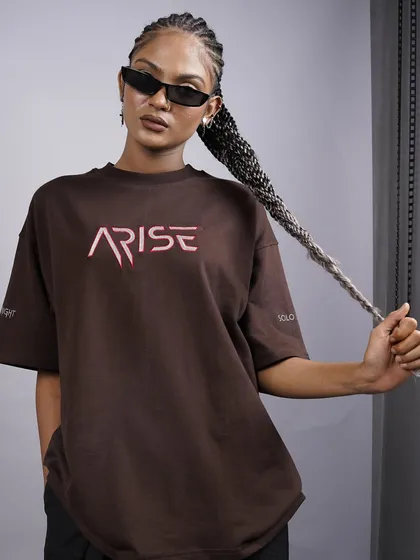 This shot is all about capturing the cool, confident attitude of modern streetwear. The clean studio background puts the focus on the model's strong pose and the graphic 'ARISE' logo on the brown oversized t-shirt, creating an image that speaks directly to a Gen-Z audience.