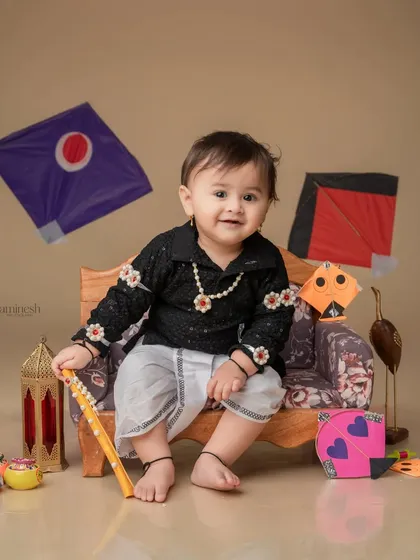 A charming portrait with floating kites in the background. The baby is seated on a miniature sofa, surrounded by festive props like lanterns and colorful firkis.