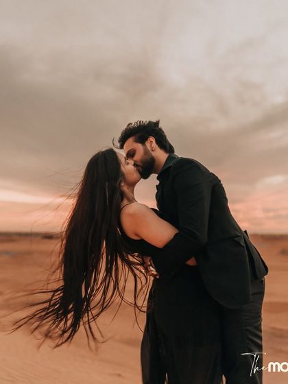 A passionate kiss in the Dubai desert as the wind catches her hair. This image is full of movement and emotion, set against the warm, dramatic colors of a desert sunset.