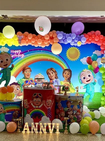 A bright and colorful Cocomelon theme birthday decoration. This setup features a large backdrop with all the main characters, a rainbow balloon arch, custom-wrapped pedestals for the cake, and a green turf floor to bring the cartoon world to life.