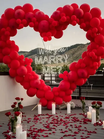 A close-up of the "Will you marry me?" neon sign inside the red balloon heart arch, surrounded by candles and rose petals for a truly romantic proposal setting.