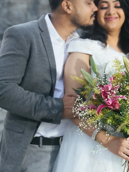 A close-up filled with warmth and affection. He leans in to kiss her neck as she smiles with her eyes closed, holding a beautiful bouquet of flowers. The focus is entirely on their intimate connection.