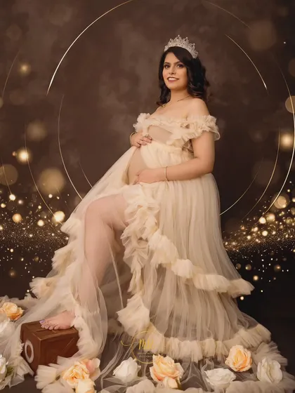 An angelic portrait of an expectant mother seated in a cream-colored ruffled gown. The scene is accented with soft lighting, a golden halo backdrop, and scattered roses for a heavenly feel.
