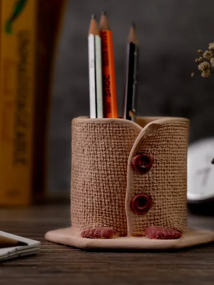 This quirky, handcrafted pen stand is styled on a wooden desk with books and dried flowers. The moody lighting and rustic setting highlight its unique texture and charming design.