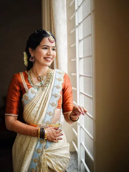 A beautiful, naturally lit portrait of a bride in a classic off-white and rust-orange silk saree. The makeup is kept soft and natural to enhance her features, creating a look of pure elegance.