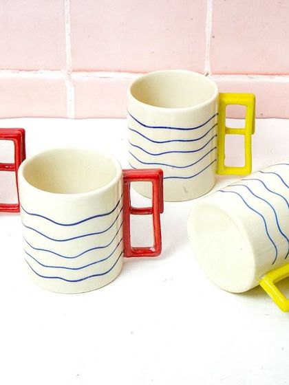 A collection of modern, artistic mugs with colorful handles. The clean, tiled background gives it a contemporary kitchen or cafe vibe.