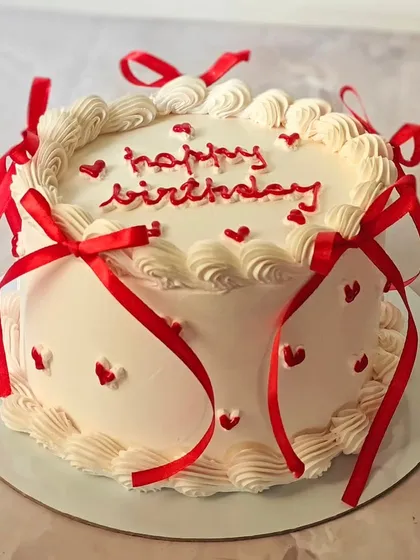 A classic white and red ribbon cake, perfect for birthdays or anniversaries. The simple design with piped borders and tiny hearts is effortlessly charming.