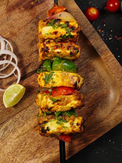A skewer of grilled paneer tikka, shot from above on a wooden tray. The char marks and fresh garnishes are captured in delicious detail.