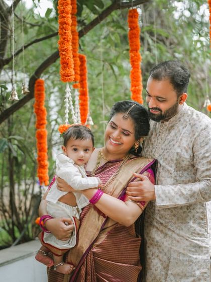 A lovely family portrait taken outdoors during a naming ceremony. The parents embrace their child under hanging marigold flower arrangements, creating a warm and traditional photograph.
