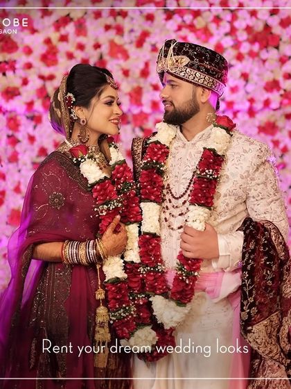 Against a wall of flowers, this couple showcases a classic wedding look. The groom's off-white sherwani is paired with a maroon stole and safa, creating a timeless and regal appearance.