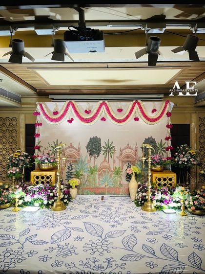 An elegant and artistic stage for an engagement. The backdrop is a printed canvas with a palace motif, framed by fresh flower garlands and traditional brass lamps.