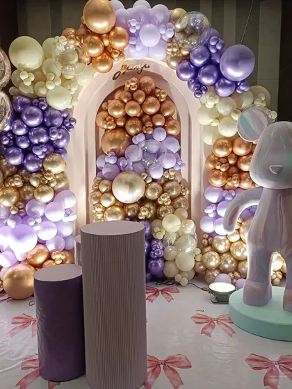 An elaborate teddy bear themed setup featuring a stunning balloon arch in metallic purple, gold, and cream. The addition of a large, stylized bear figure and matching pedestals elevates the theme into a luxurious and modern celebration.