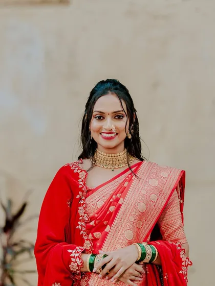 A portrait of a smiling bride in her red saree. The half-up hairstyle with soft tendrils frames her face beautifully, creating a romantic and elegant look.