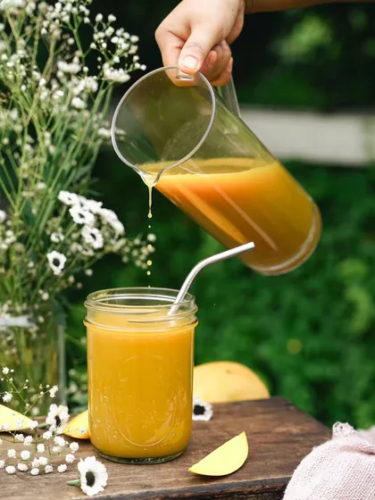 A perfect summer scene with a pitcher of fresh mango juice being poured into a glass. The outdoor setting with natural greenery makes it feel like a lovely garden picnic.
