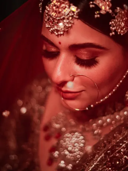 A close-up bridal portrait under a red veil. The lighting highlights her beautiful makeup and traditional jewelry, creating a dramatic and intimate feel.
