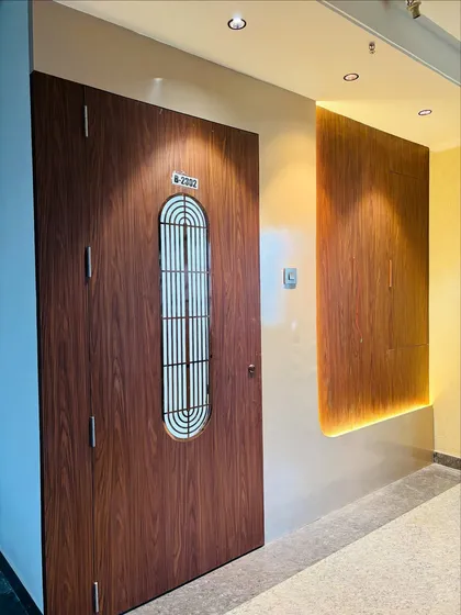 A modern wooden safety door with a vertical grill, complemented by a backlit wooden panel on the adjacent wall for a warm, ambient glow.
