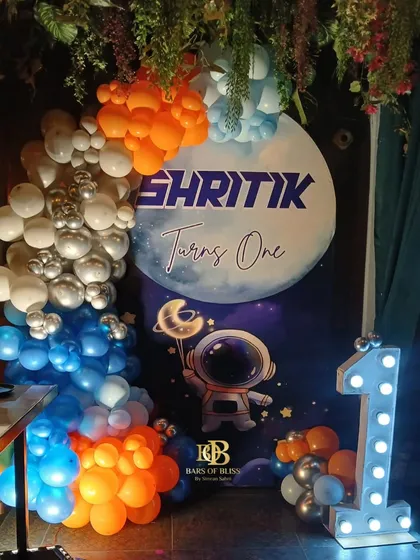 A space-themed first birthday party for a little astronaut. The decor features a personalized backdrop with a moon and astronaut graphic, framed by balloons in orange, blue, and silver, and a light-up number '1'.